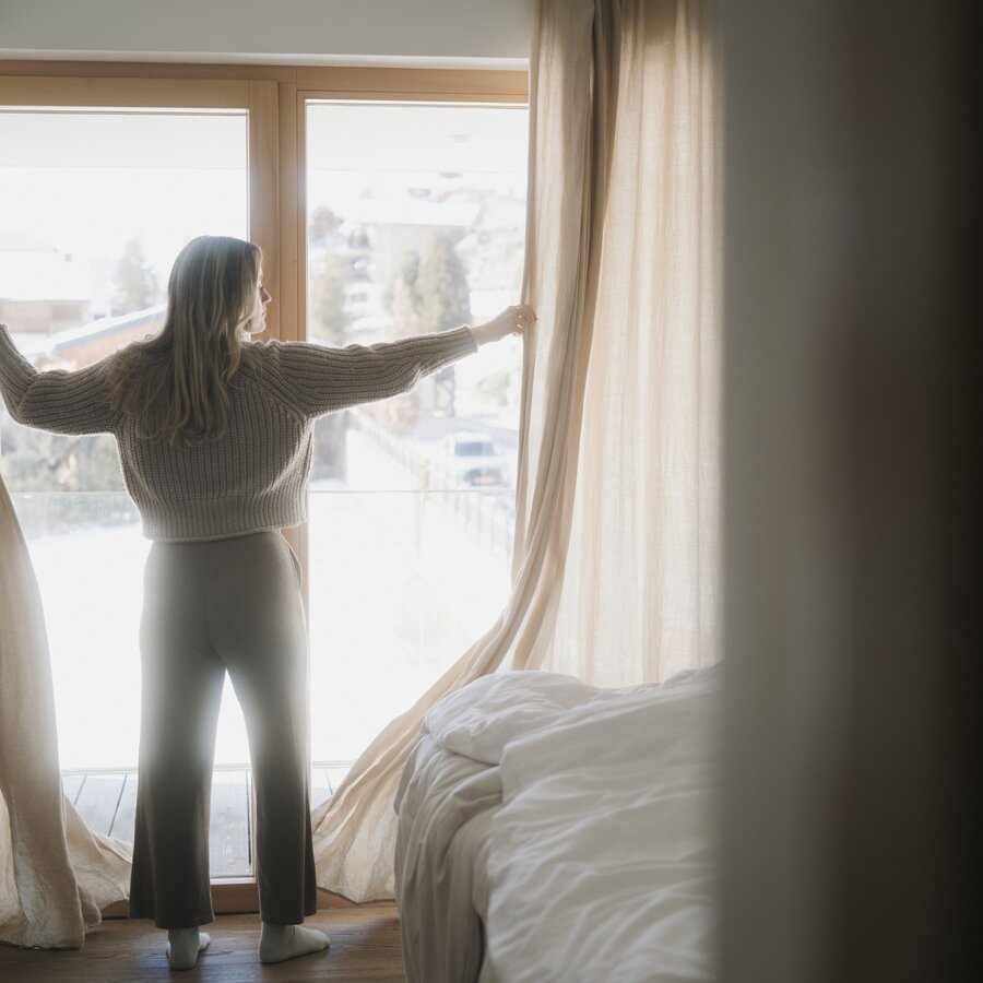 Woman standing by large window, drawing back beige curtains