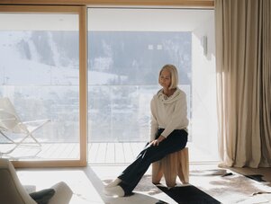Older woman sits on wooden stool by glass door, snow outside.